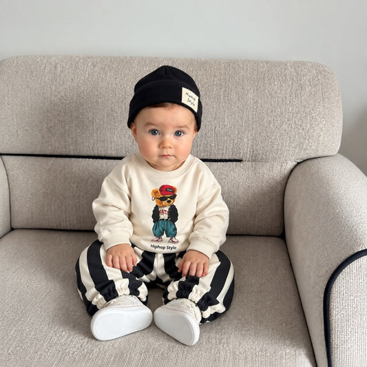 Baby wearing a cream bear sweatshirt, black striped joggers and black beanie
