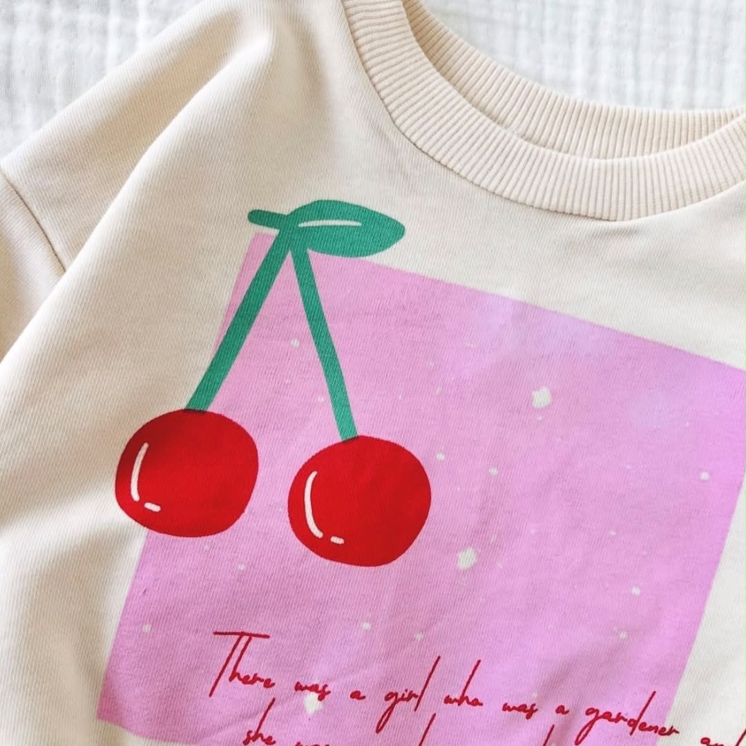 Close-up of red cherry graphic and pink panel on cream baby sweatshirt