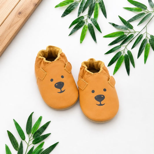 Baby Bear Face Mustard Yellow Leather Soft-Sole Moccasins flat lay with green leaves