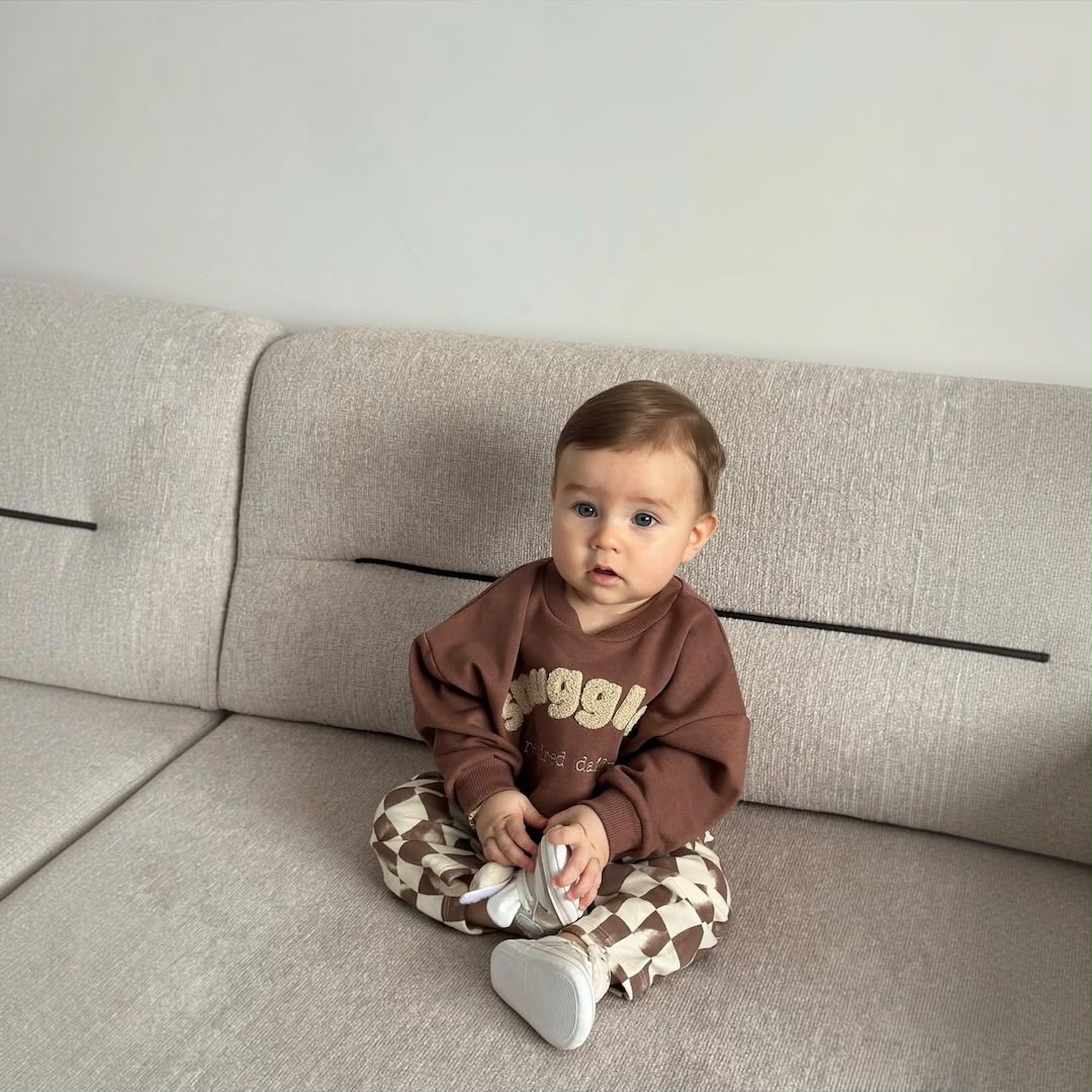 Baby wearing a brown sweatshirt and checkered pants while sitting