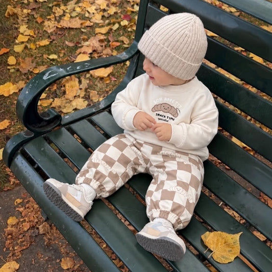 Baby wearing croissant sweatshirt and brown checkered joggers on an outdoor bench